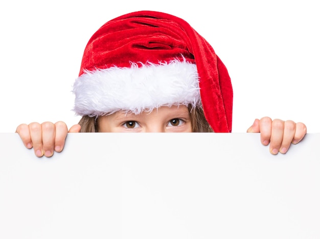 Foto menina vestindo um chapéu de papai noel posando atrás de um painel branco isolado em fundo branco criança segurando um outdoor de natal vazio criança espreitando atrás de um quadro de feriados em branco