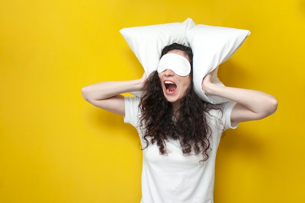 Foto menina zangada com máscara de sono e pijama com almofadas gritando em fundo amarelo