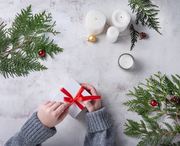 Meninas abrindo o presente de Natal branco com fita vermelha na mesa cinza claro. Fundo de Natal com árvore de thuja, velas e bolas de Natal. Camada plana e vista superior