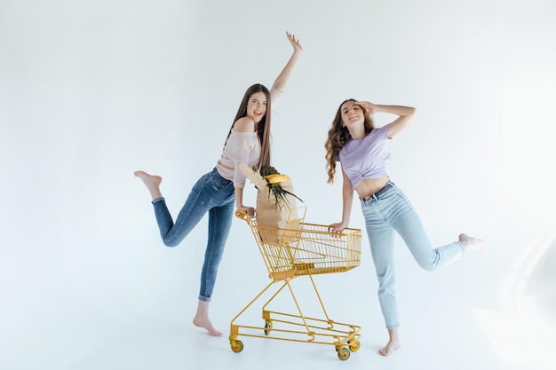 meninas animadas com saco de compras sentado no carrinho de compras, isolado no branco