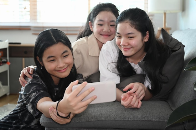 Meninas asiáticas alegres tomando selfie com smartphone inteligente enquanto sentam juntas na sala de estar