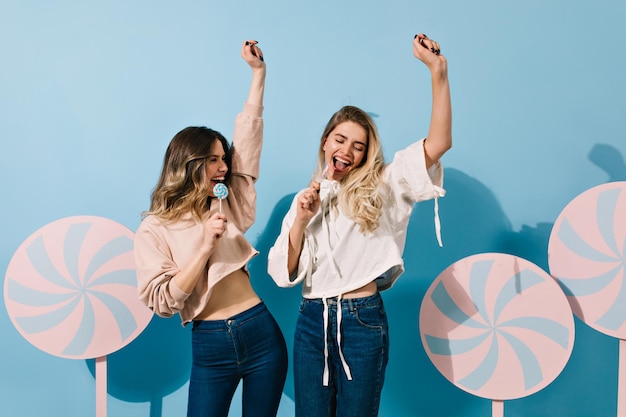 Meninas bem-humoradas com pirulitos dançando em fundo azul senhoras encantadoras lambendo doces