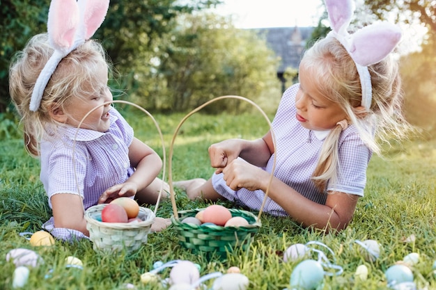 Meninas bonitas em orelhas de coelho engraçadas jogam em um jogo coletando ovos de Páscoa em uma cesta de vime em uma grama contra fundo verde desfocado Conceito de férias de primavera