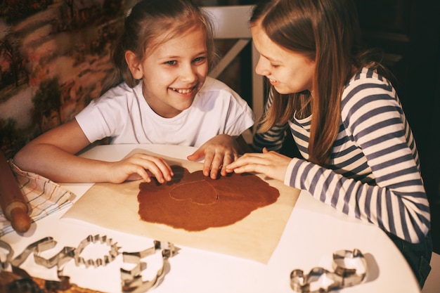 Meninas bonitas na cozinha de casa à mesa cortam biscoitos em forma de coração da massa