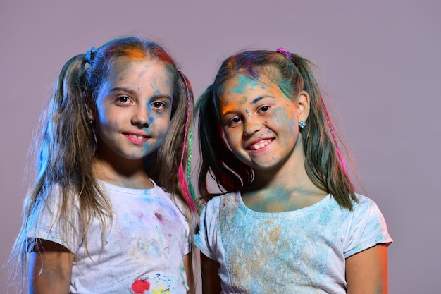 Meninas com rostos sorridentes ficam no fundo roxo, copie o espaço. Conceito de criatividade e educação. Crianças com rabos de cavalo ficam perto. As alunas têm manchas de tinta nos rostos e nas camisetas.