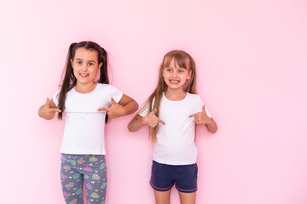 Meninas de crianças espantadas mostrando t-shirt branca, isolada no fundo rosa. conceito de estilo de vida da infância. mock up espaço de cópia. apontando o dedo indicador de lado