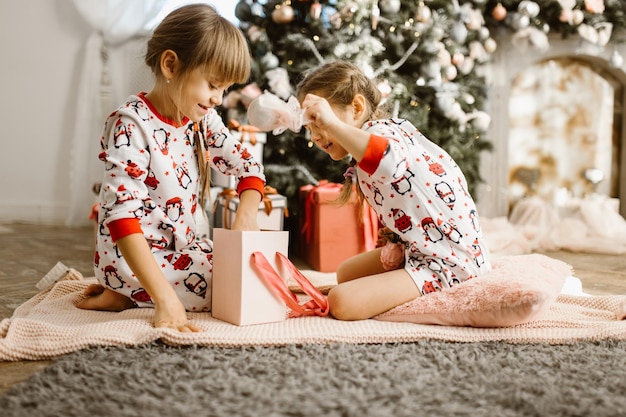 Foto meninas de pijama abrem a caixa de presente debaixo da árvore de natal