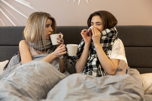 Meninas europeias insalubres, deitado na cama, debaixo de cobertores e bebendo chá quente em xícaras. Mulheres jovens usam lenços. Uma garota espirrando em um lenço de papel. Interior do quarto em apartamento moderno