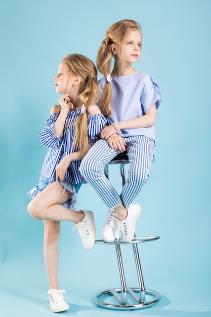 Meninas gêmeas em roupas azuis claras estão posando perto de um banquinho em um azul.