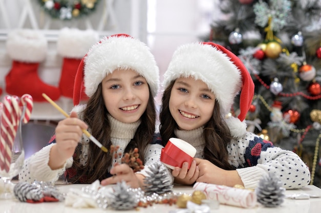 Meninas gêmeas felizes em chapéus de Papai Noel se preparando para o Natal sentadas à mesa olhando para a câmera