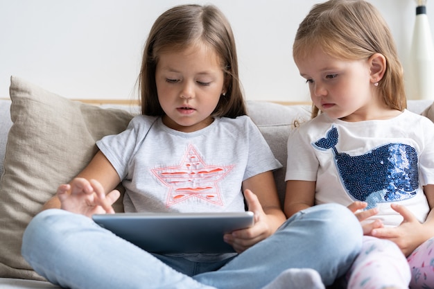 Meninas ou irmãs sentadas no sofá jogando tablet digital na sala de estar em casa.