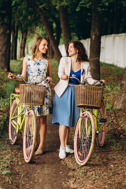 Meninas positivas e felizes que andam com as bicicletas no parque do beco, dia de verão. Amigos do sexo feminino desfrutando de um passeio na rua com suas bicicletas.