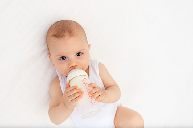 Menino 8 meses de idade, deitado na cama no berçário de costas e segurando uma garrafa de leite, alimentando o bebê, conceito de comida para bebê