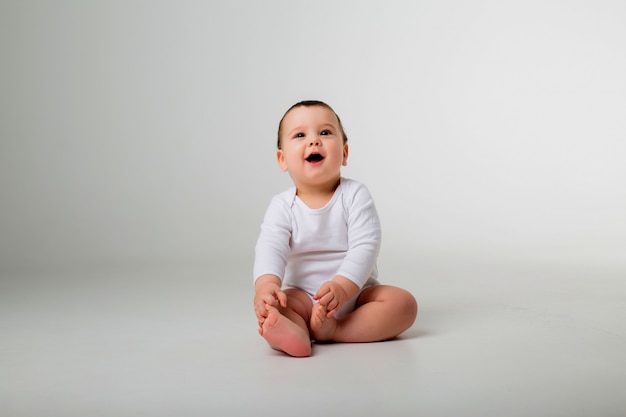 Foto menino 9 meses em uma roupa branca, sentado em uma parede branca