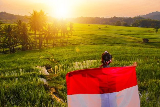 Foto menino acenando bandeira vermelha e branca indonésia com vista para os campos de arroz à tarde