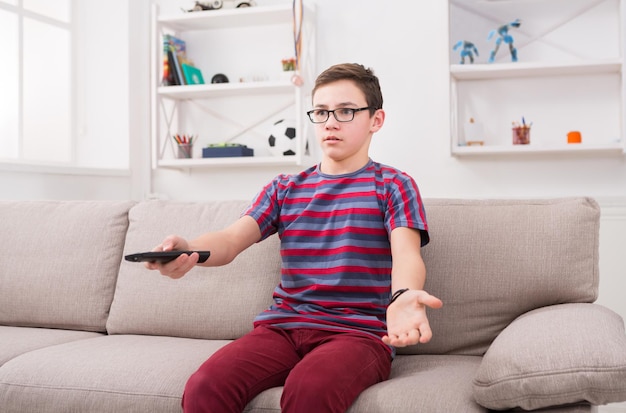 Menino adolescente assistindo televisão, usando o controle remoto para mudar de canal. Jovem insatisfeito com o que vê na tela da TV. Sentado no sofá na sala de estar de casa