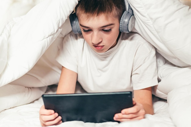 Menino adolescente deitado na cama sob um cobertor branco aprende a tocar em um tablet, computador ou telefone ouve música em fones de ouvido