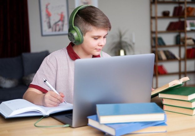 Menino adolescente em fones de ouvido, estudando online usando laptop, escrevendo no caderno, lendo o livro.