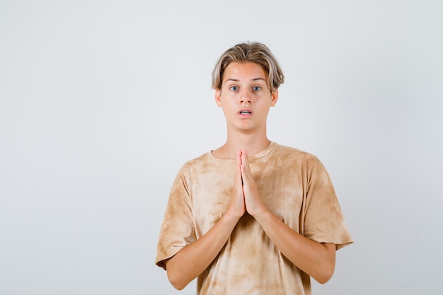 Menino adolescente em t-shirt, mantendo as mãos em gesto de oração e olhando perplexo, vista frontal.