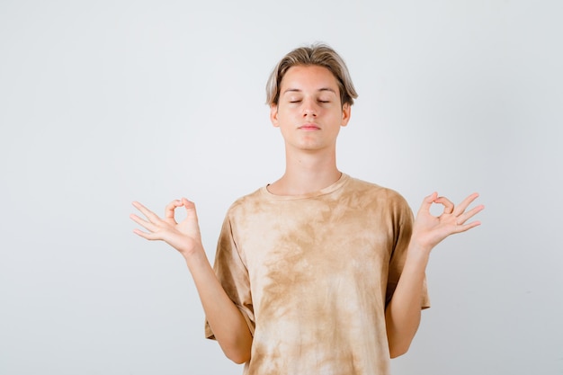 Menino adolescente em t-shirt mostrando sinal de mudra, mantendo os olhos fechados e olhando relaxado, vista frontal.