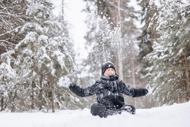 Menino adolescente feliz sentado na neve na floresta de inverno. criança se divertindo ao ar livre. alegre adolescente jogando na neve na queda de neve. rindo criança sorridente andando em winter park em tempo frio