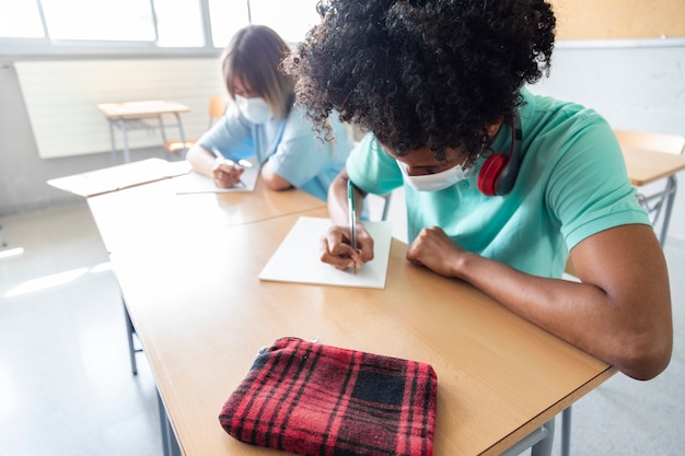 Foto menino adolescente negro usando máscara facial na classe estudante do ensino médio escrevendo espaço para cópia educação