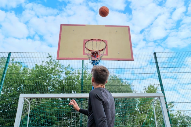 Menino adolescente pulando com a bola jogando basquete de rua, vista traseira. estilo de vida ativo e saudável, hobbies e lazer, conceito de juventude