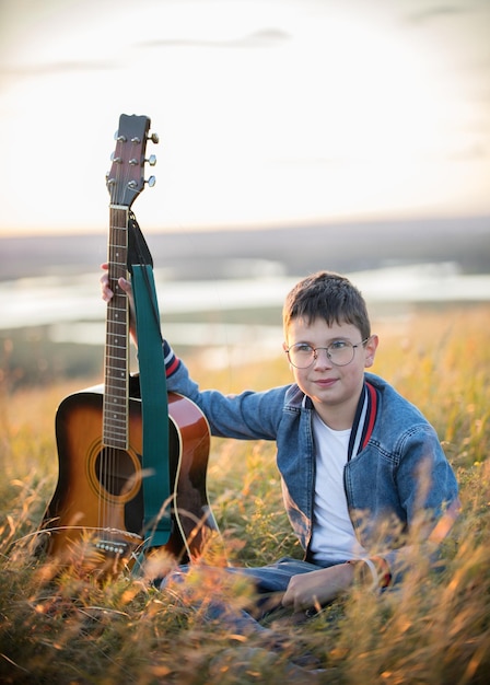 Menino adolescente tocando violão no campo de verão no rosto sorridente do sol, olhando para a câmera
