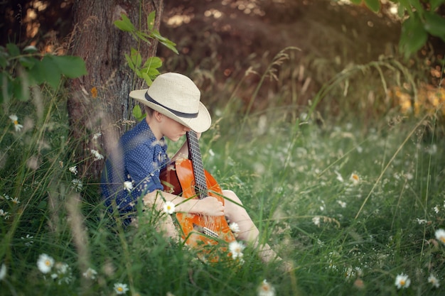 Menino adorável com a guitarra, relaxando no parque. Garoto sentado em uma grama no dia de verão