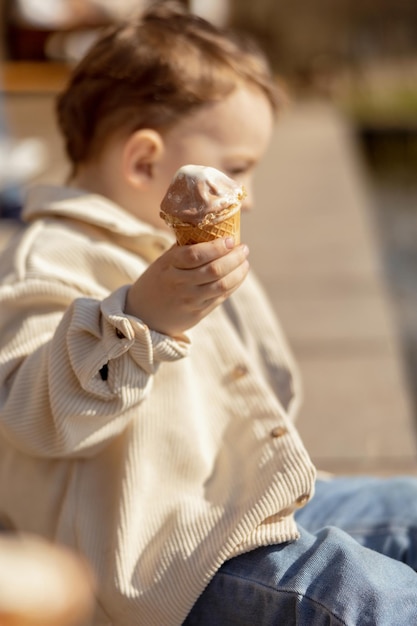 Menino adorável sentado ao ar livre e comendo sorvete Água do lago e clima ensolarado Criança e doces Açúcar Criança desfruta de uma deliciosa sobremesa Criança pré-escolar com roupas casuais Emoção positiva