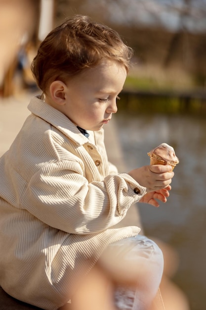 Menino adorável sentado ao ar livre e comendo sorvete Água do lago e clima ensolarado Criança e doces Açúcar Criança desfruta de uma deliciosa sobremesa Criança pré-escolar com roupas casuais Emoção positiva