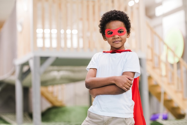 Menino africano fofo em trajes casuais brancos e manto vermelho de super-homem olhando para você enquanto brinca no jardim de infância