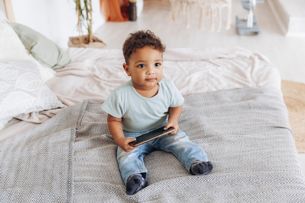 Menino afro-americano bonitinho com smartphone em casa na cama criança feliz assistindo desenhos no telefone