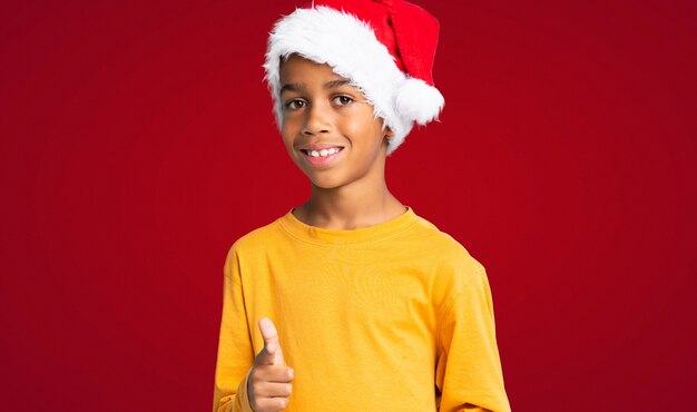 Menino afro-americano com chapéu de Natal aponta o dedo para você com uma expressão confiante sobre fundo vermelho