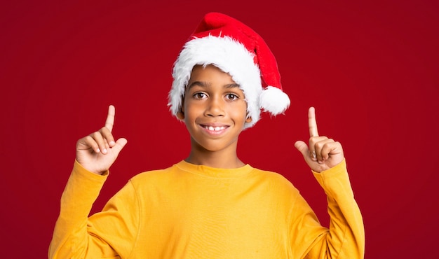 Menino afro-americano com chapéu de natal apontando uma ótima idéia sobre fundo vermelho