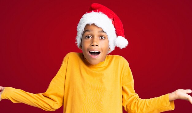 Menino afro-americano com chapéu de Natal com expressão facial chocado sobre parede vermelha