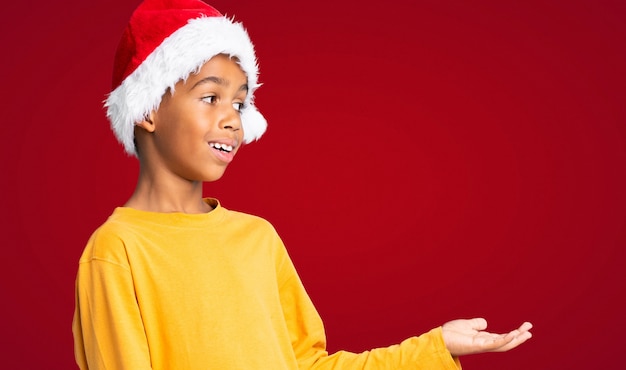 Menino afro-americano com chapéu de natal com expressão facial de surpresa sobre parede vermelha