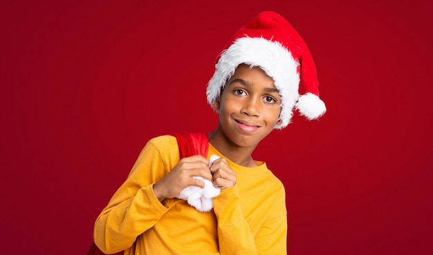 Menino afro-americano com chapéu de Natal e levar uma sacola com presentes ao longo da parede vermelha