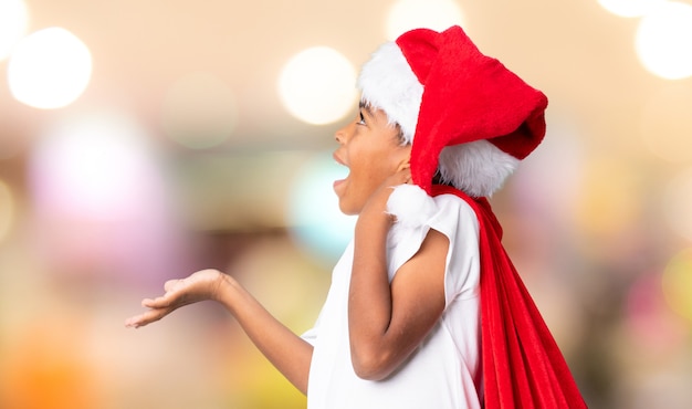 Menino afro-americano com chapéu de Natal e levar uma sacola com presentes, fazendo o gesto de surpresa sobre parede desfocada