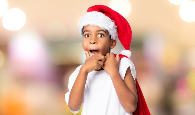 Menino afro-americano com chapéu de Natal e levar uma sacola com presentes sobre fundo desfocado