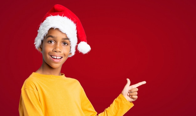 Menino afro-americano com chapéu de natal surpreso e apontando o lado ao longo da parede vermelha