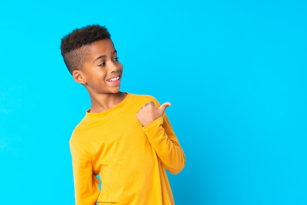 Menino afro-americano sobre azul isolado apontando para o lado para apresentar um produto