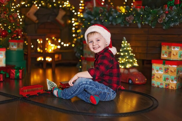 Menino alegre com chapéu de papai noel brincando com a locomotiva em casa enquanto está sentado perto da árvore de natal