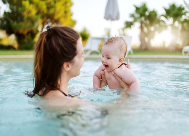 Menino alegre de 6 meses, caucasiano, aprendendo a nadar na piscina. Mãe segurando seu filho. Primeira vez no conceito de piscina.