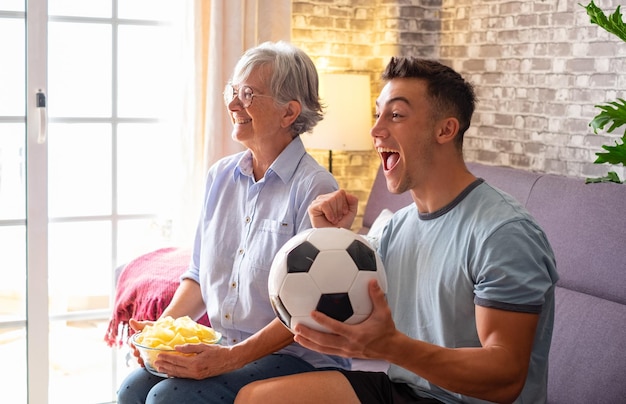 Menino alegre e fãs de futebol da avó sênior assistindo a um jogo de futebol na tv sentado em um sofá confortável na sala de estar