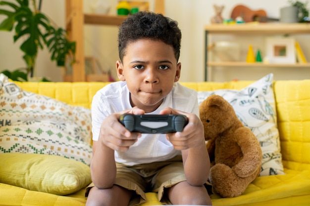 Foto menino alegre e fofo de etnia africana com joystick olhando para você com um sorriso enquanto joga videogame no sofá enquanto se diverte