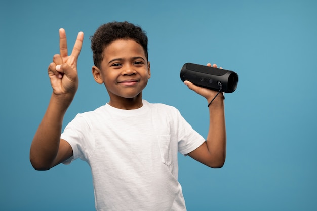 Menino alegre e fofo de origem africana mostrando um gesto de paz enquanto se diverte e ouve música na coluna smart wireless