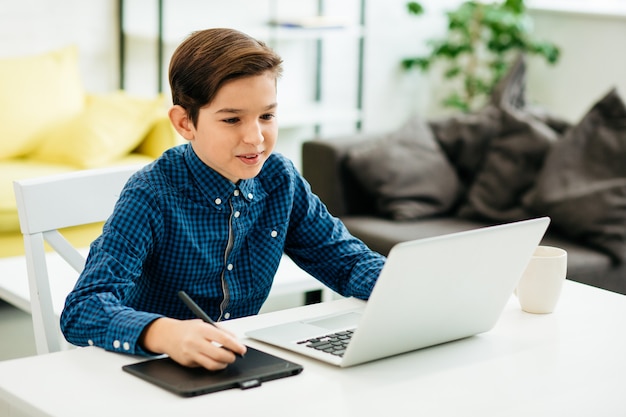 Menino alegre e inteligente sentado à mesa com um laptop moderno na frente dele e sorrindo enquanto coloca a caneta na almofada de desenho