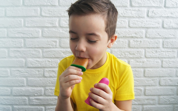 Menino alegre e sorridente comendo sorvete de gelo suculento no fundo da parede de tijolos