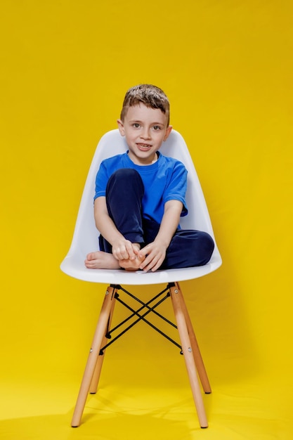 Menino alegre em uma camiseta casual azul e calças sentadas em uma cadeira branca no estúdio de fundo amarelo